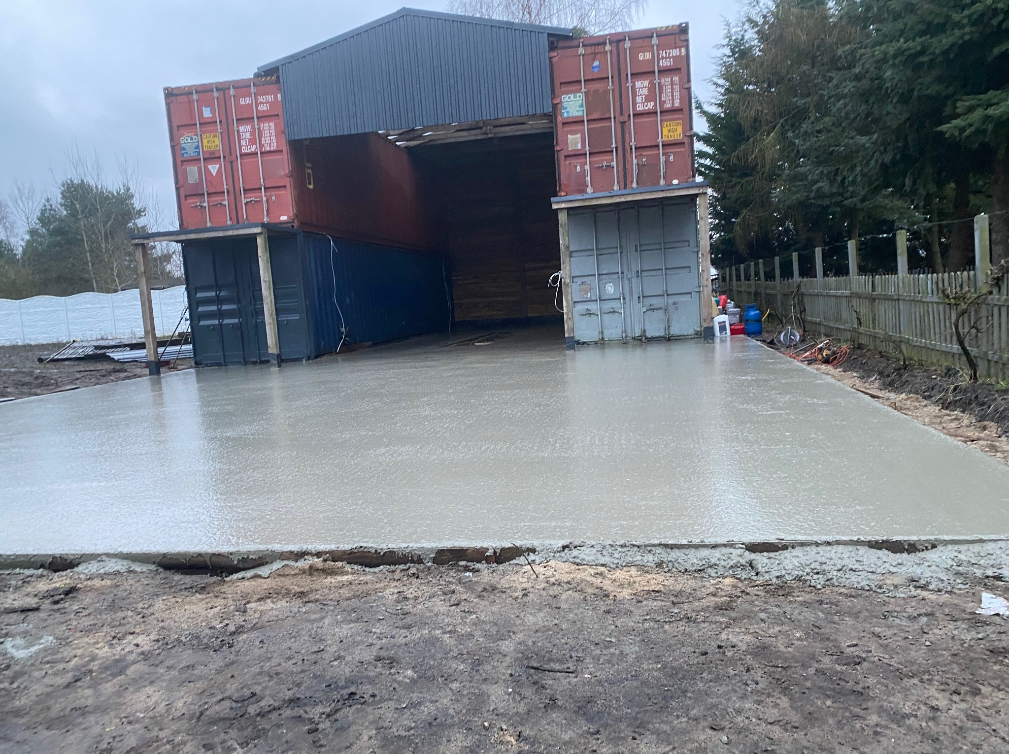 Wylany, świeży beton na przygotowanym podłożu, z konstrukcją z kontenerów morskich w tle, tworzącą zadaszenie. Widoczne elementy szalunku i otaczający teren.