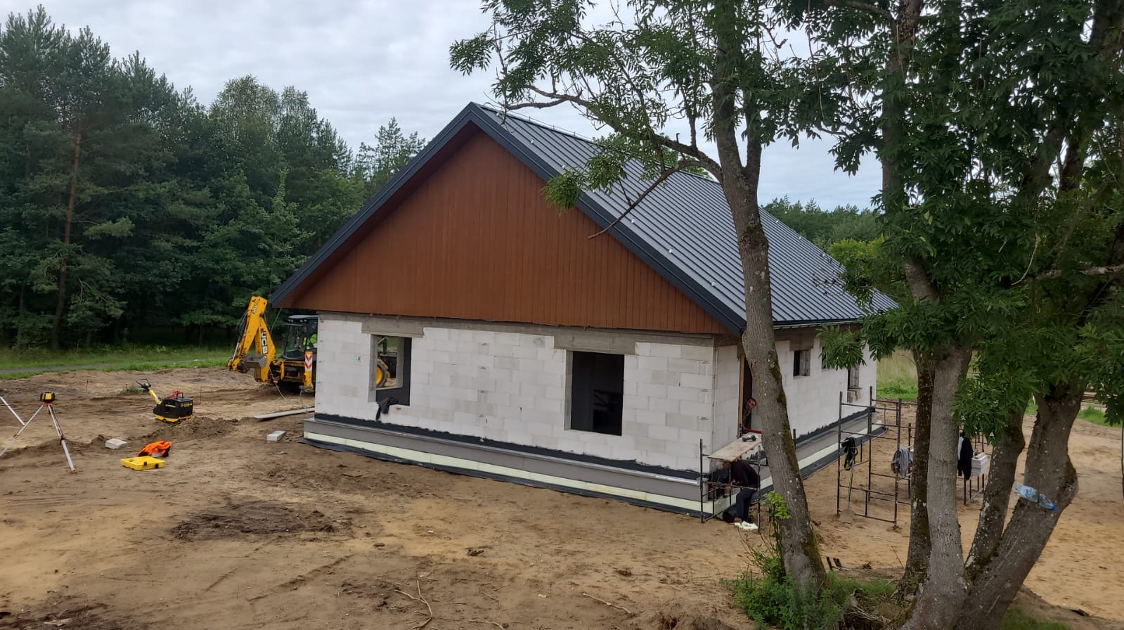 Surowy stan domu jednorodzinnego z białego bloczka, z brązową deską pod dachem i ciemnoszarą blachą, widoczny sprzęt budowlany i drzewa w tle.