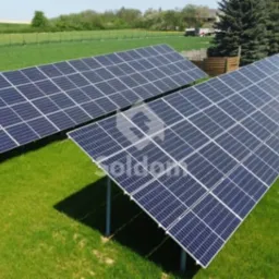 Dwa rzędy paneli słonecznych ustawione na zielonej trawie z widocznym ogrodzeniem i drzewami w tle.