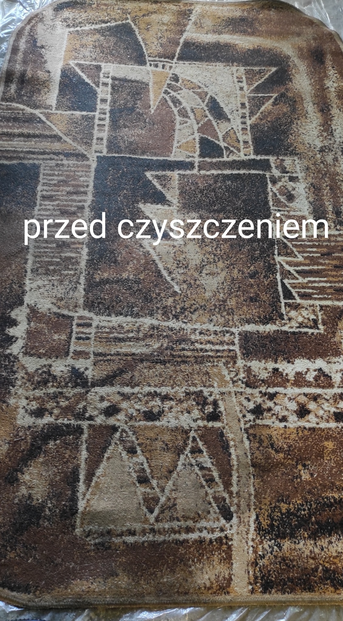 Brudny dywan z geometrycznym wzorem w odcieniach brązu i beżu, sfotografowany z góry, z widocznym napisem 'przed czyszczeniem'.
