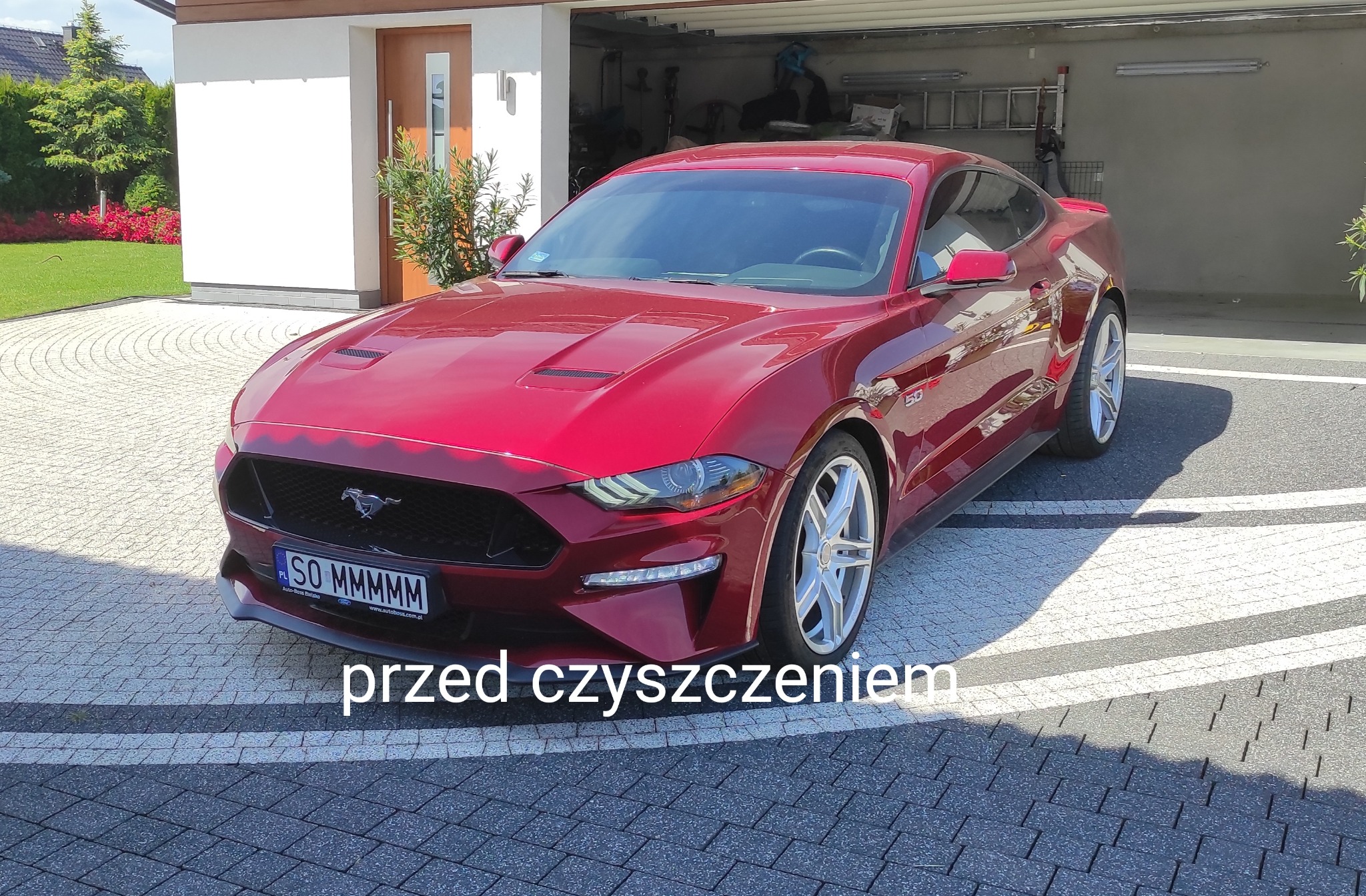 Czerwony Ford Mustang zaparkowany na kostce brukowej przed garażem, widoczny numer rejestracyjny SO MMMMM.
