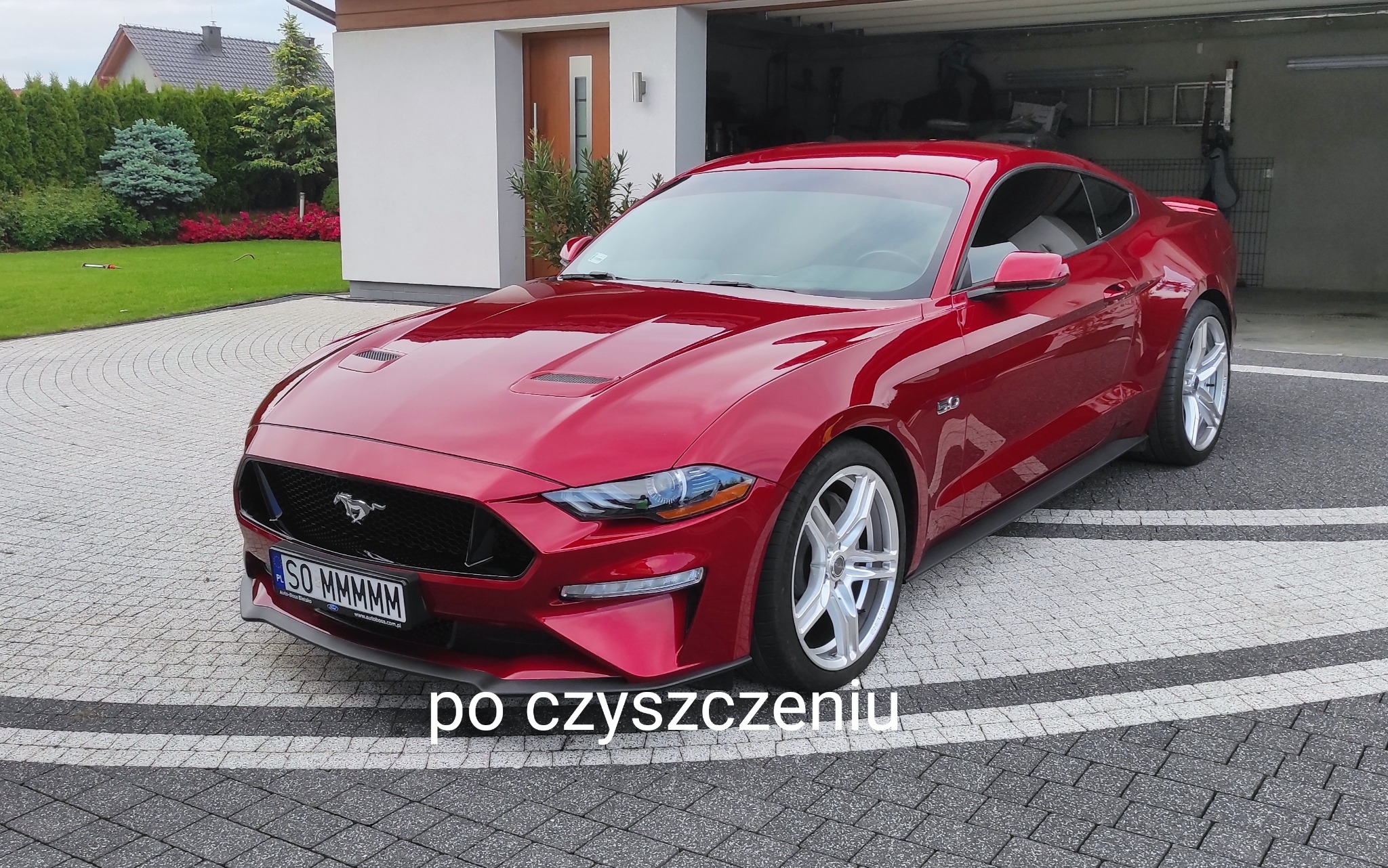 Czerwony samochód sportowy Ford Mustang zaparkowany na kostce brukowej przed garażem, widoczne detale karoserii po czyszczeniu, tablica rejestracyjna SO MMMMMM.