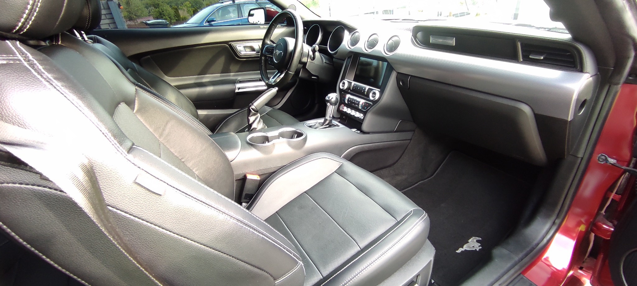 Wnętrze samochodu sportowego z czarną skórzaną tapicerką, srebrnymi akcentami na desce rozdzielczej i logo na dywaniku, widok z otwartych drzwi.