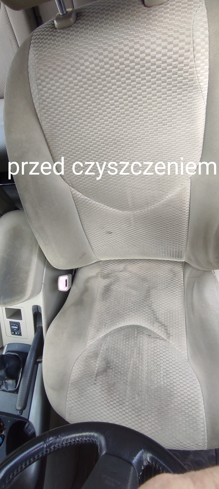 Tapicerka fotela samochodowego z widocznymi plamami i zabrudzeniami przed procesem czyszczenia, widok z góry na fotel kierowcy, z fragmentem kierownicy w dolnej części kadru.