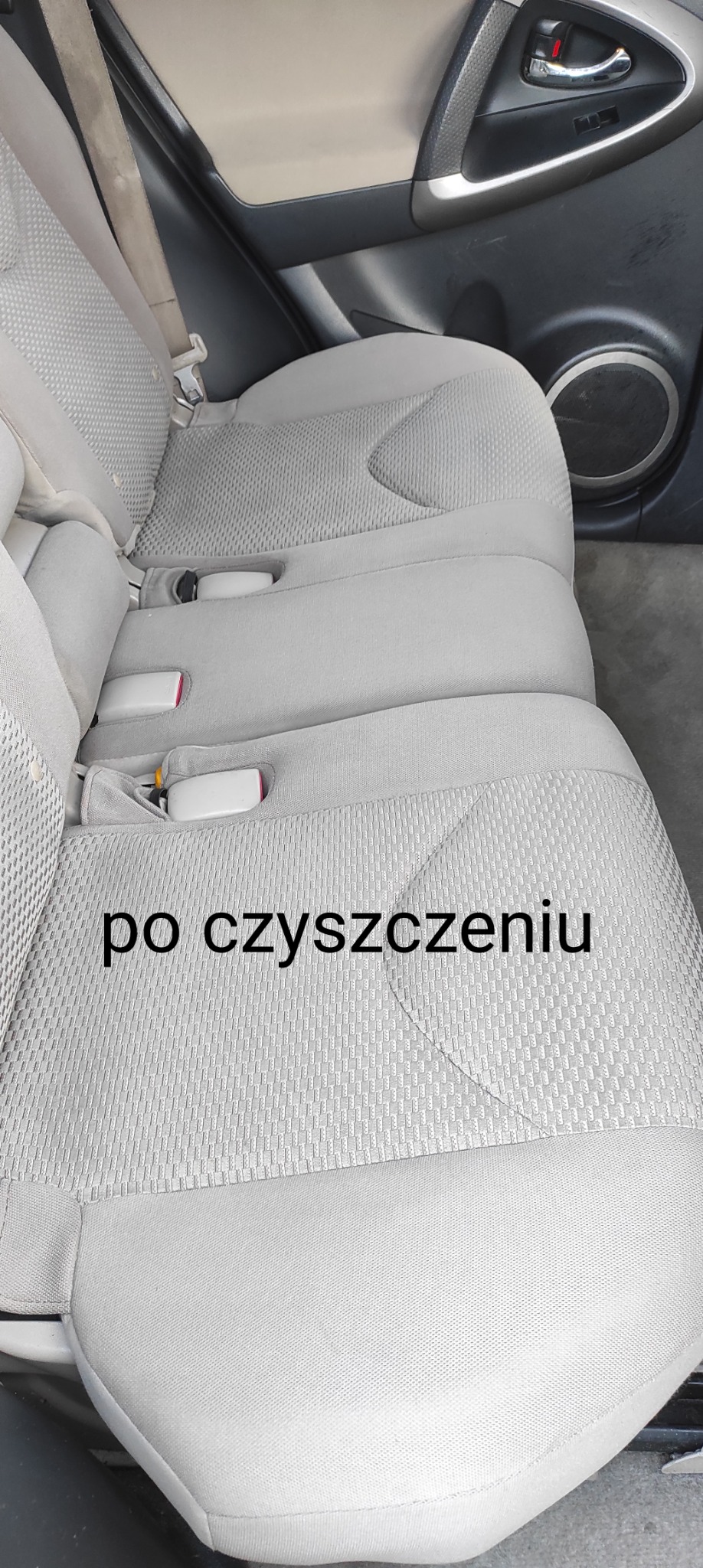 Jasnoszara, tekstylna tapicerka tylnej kanapy samochodu po czyszczeniu, widoczne pasy bezpieczeństwa i fragment drzwi z głośnikiem.