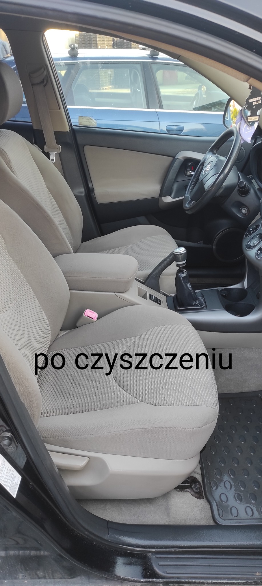 Wnętrze czarnego samochodu Toyota RAV4 po czyszczeniu tapicerki, widoczne jasne, odświeżone fotele z tkaniny w neutralnym odcieniu beżu, czarna kierownica i deska rozdzielcza, gumowe dywaniki...