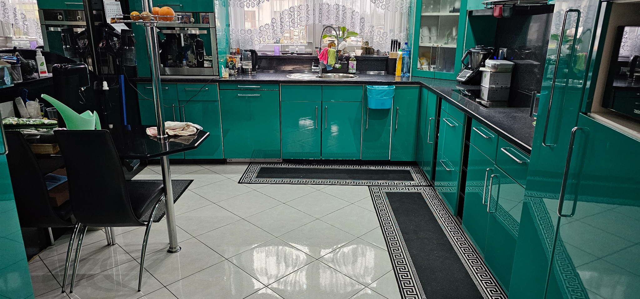 Kuchnia w odcieniach turkusu z czarnymi blatami i białymi płytkami podłogowymi, widoczne akcesoria kuchenne i dywaniki z motywem greckim.