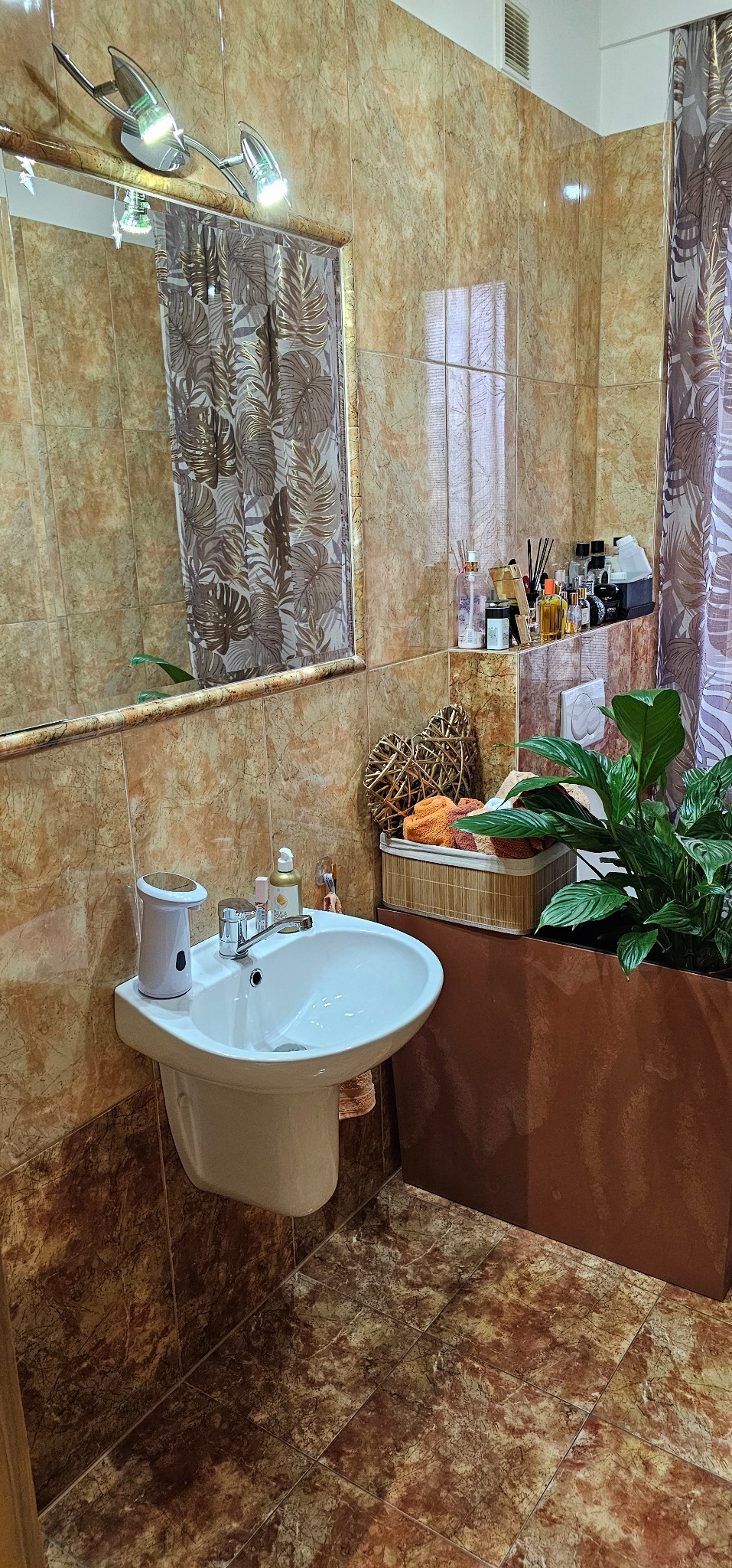 Umywalka ceramiczna z automatycznym dozownikiem mydła w łazience wyłożonej marmurkowymi płytkami, z dekoracyjnym lustrem i roślinami doniczkowymi.