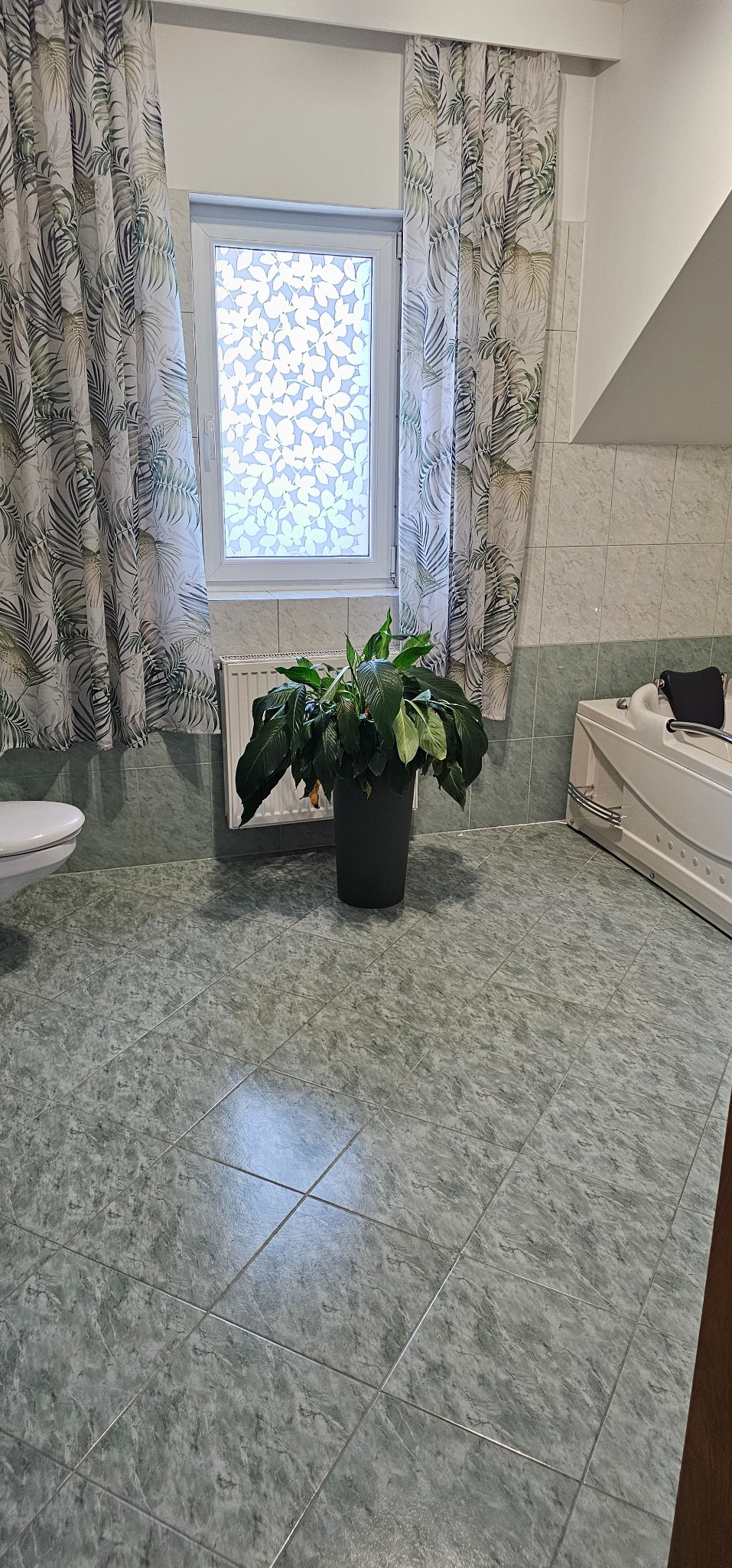 Jasna łazienka z zielonymi płytkami podłogowymi, oknem z mleczną folią, zasłonami w liście, białą wanną z hydromasażem i doniczką z zieloną rośliną.