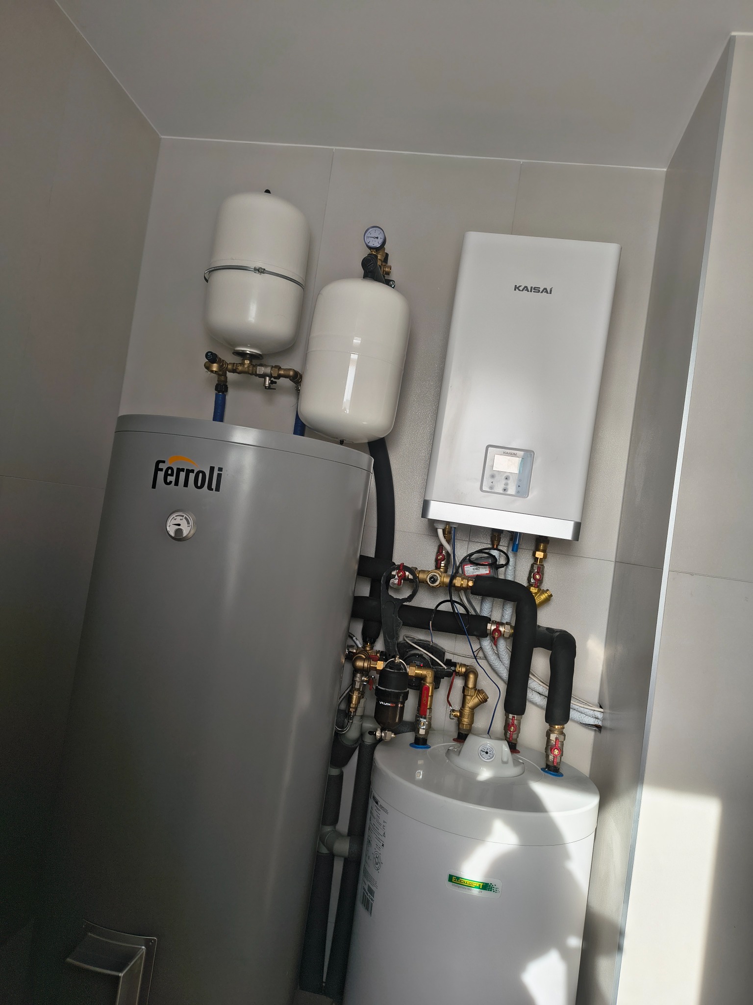 Zestaw urządzeń grzewczych: szary zasobnik Ferroli, biały piec Kaisai i dwa białe zbiorniki wyrównawcze, połączone rurami w schludnej kotłowni.