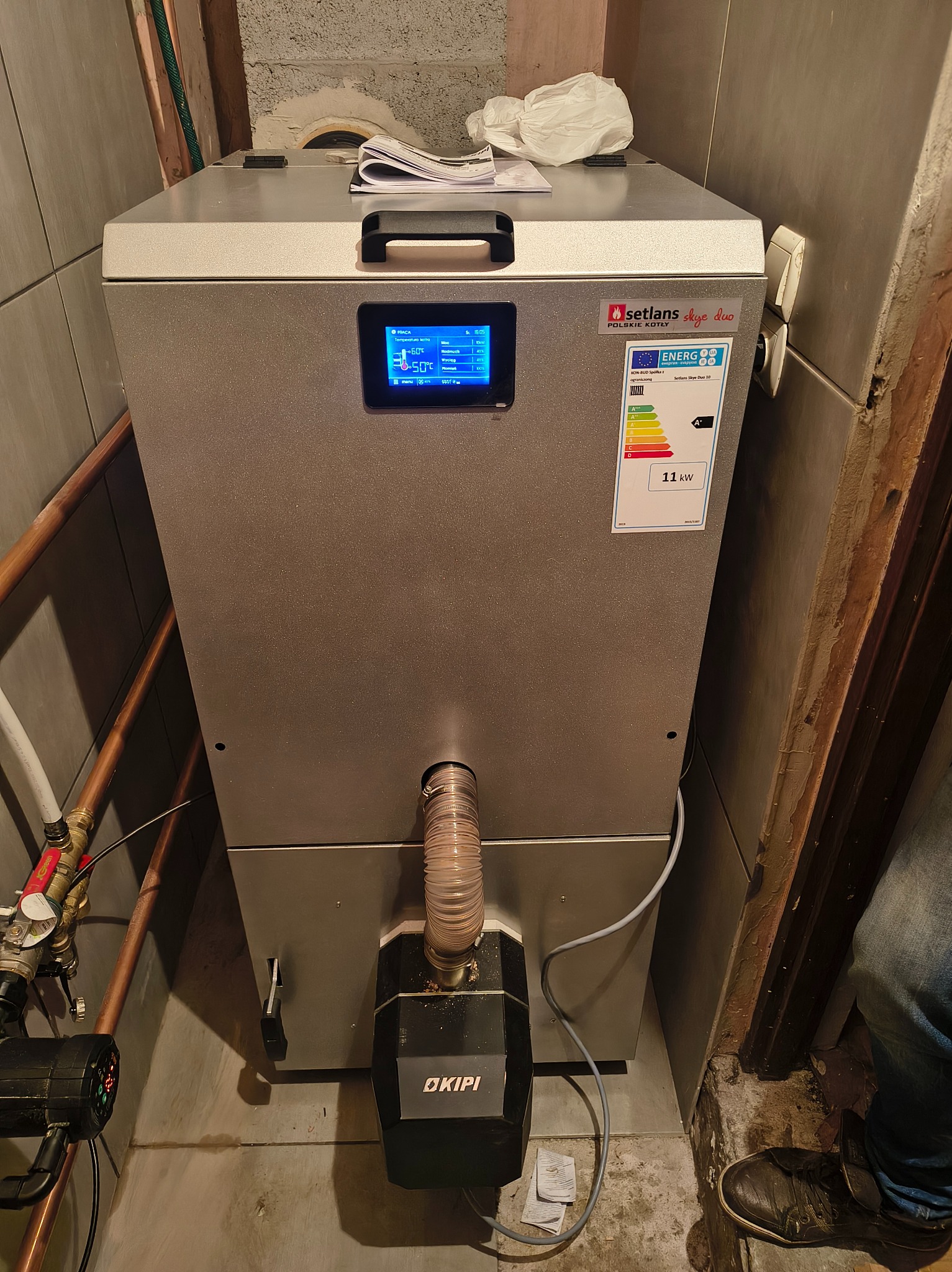 Szary piec CO marki Setlans Skye Duo 10 o mocy 11kW z etykietą energetyczną A+ i wyświetlaczem, zamontowany w kotłowni. Widoczne rury i elementy instalacji.