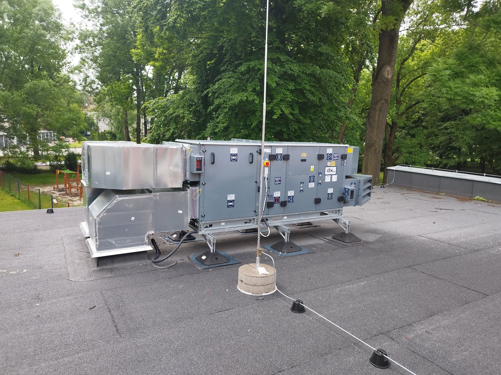 Szara centrala wentylacyjna na dachu budynku, z widocznymi przewodami i elementami montażowymi, na tle zieleni drzew. Widok z zewnątrz, w dzień.