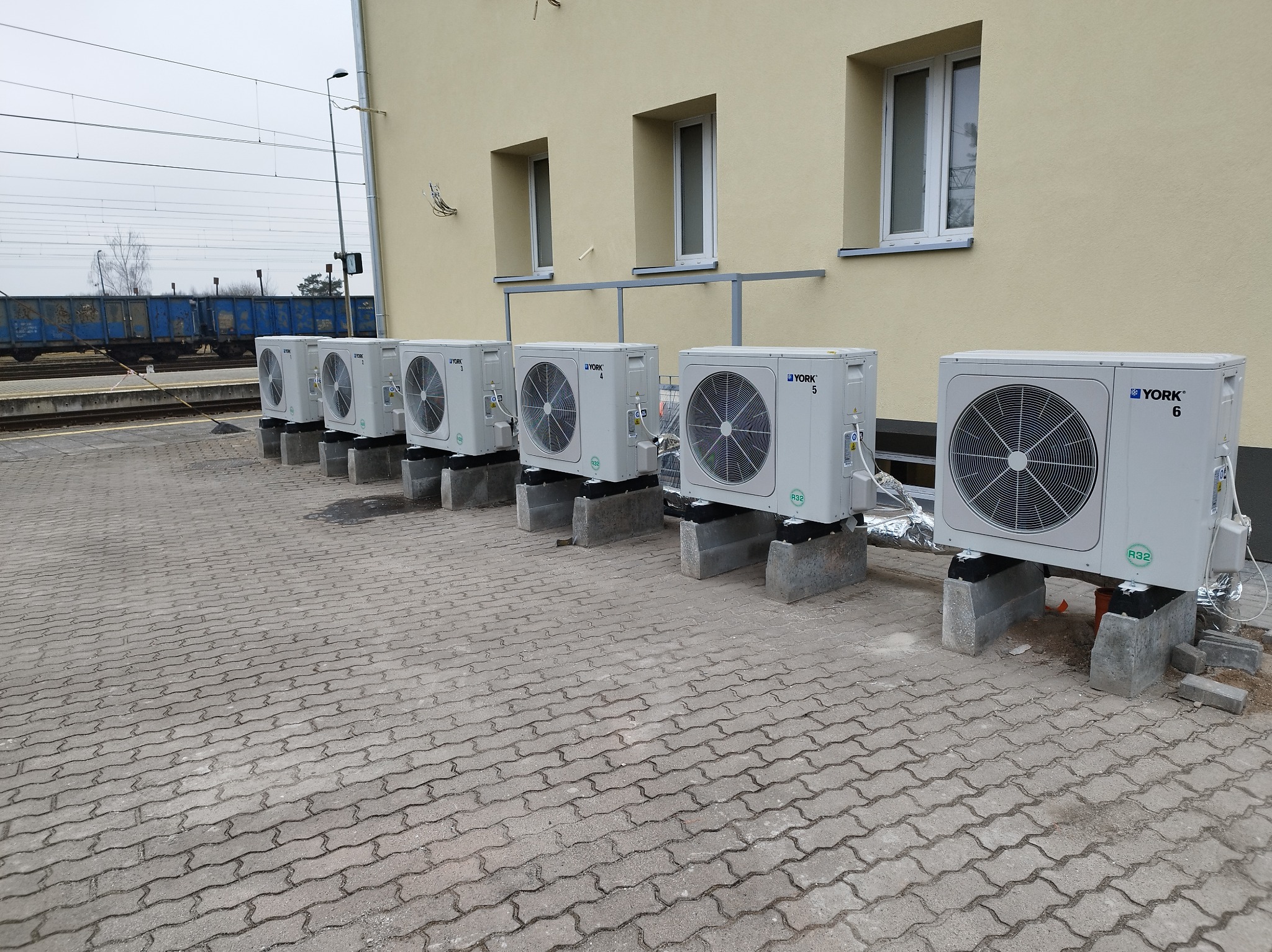 Rząd klimatyzatorów York na betonowych podstawach, ustawionych wzdłuż ściany budynku. W tle widoczny wagon kolejowy i szyny. Instalacja zewnętrzna, widok z dołu.