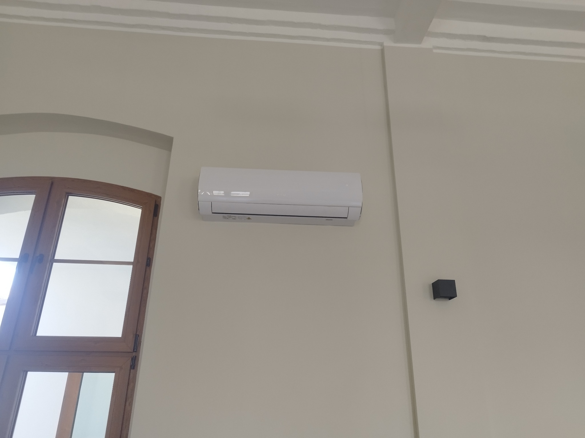 Biały klimatyzator ścienny zamontowany w jasnym pomieszczeniu obok okna z drewnianą ramą i czujnika. Minimalistyczny design, neutralne tło.