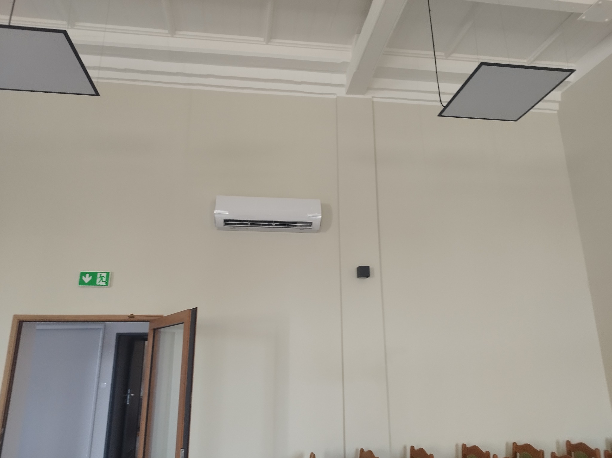 Biały klimatyzator ścienny w jasnym pomieszczeniu z otwartymi drzwiami i wiszącymi lampami sufitowymi. Widoczny znak ewakuacyjny.