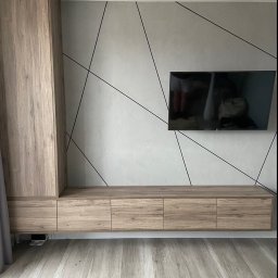 Dmytro Kostin REMONT-PROF - Drewniana zabudowa ściany z szafkami i szafą, telewizor na ścianie z geometrycznym wzorem z czarnych linii, podłoga z paneli w odcieniach szarości i beżu, dwa białe reflektory sufitowe.