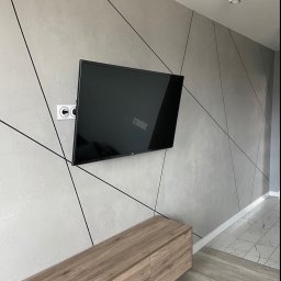 Dmytro Kostin REMONT-PROF - Szara ściana z dekoracyjną strukturą i czarnymi liniami tworzącymi geometryczny wzór, wiszący telewizor, drewniana szafka RTV i białe gniazdko elektryczne.