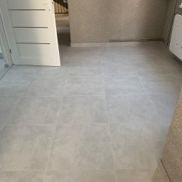 Dmytro Kostin REMONT-PROF - Jasnoszare płytki podłogowe ułożone w kwadraty z wąskimi fugami, widoczna biała ściana z grzejnikiem, czerwona gaśnica, fragment otwartych białych drzwi z poziomymi wgłębieniami i schody w tle.