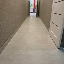 Dmytro Kostin REMONT-PROF - Korytarz po remoncie z jasnymi płytkami podłogowymi, ściany wykończone strukturalnym tynkiem w odcieniach brązu, białe drzwi po prawej i widok na pokój z pomarańczową zasłoną w tle.