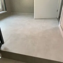 Jasne, kwadratowe płytki podłogowe ułożone w pomieszczeniu z szarą, teksturowaną ścianą i białymi drzwiami.