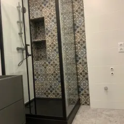 Nowoczesna kabina prysznicowa z czarną ramą, wyłożona płytkami w geometryczne wzory, z wnęką na kosmetyki, sąsiadująca z białą ścianą z poziomymi panelami i gniazdkiem elektrycznym.