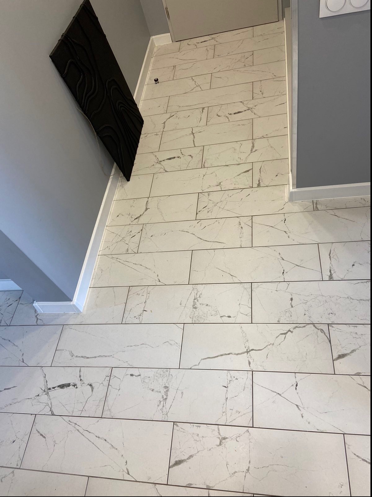 Ułożona podłużna, biała płytka podłogowa z szarym marmurkowym wzorem, widok z góry, dekoracyjna czarna płaskorzeźba na szarej ścianie, białe listwy przypodłogowe.