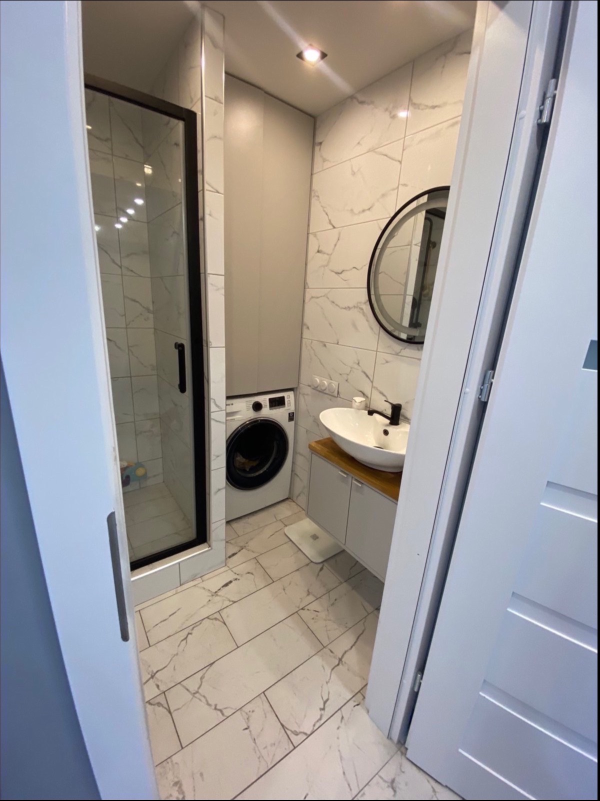 Nowoczesna łazienka z kabiną prysznicową, pralką pod szafką i umywalką nablatową z okrągłym lustrem, wykończona białymi płytkami imitującymi marmur.