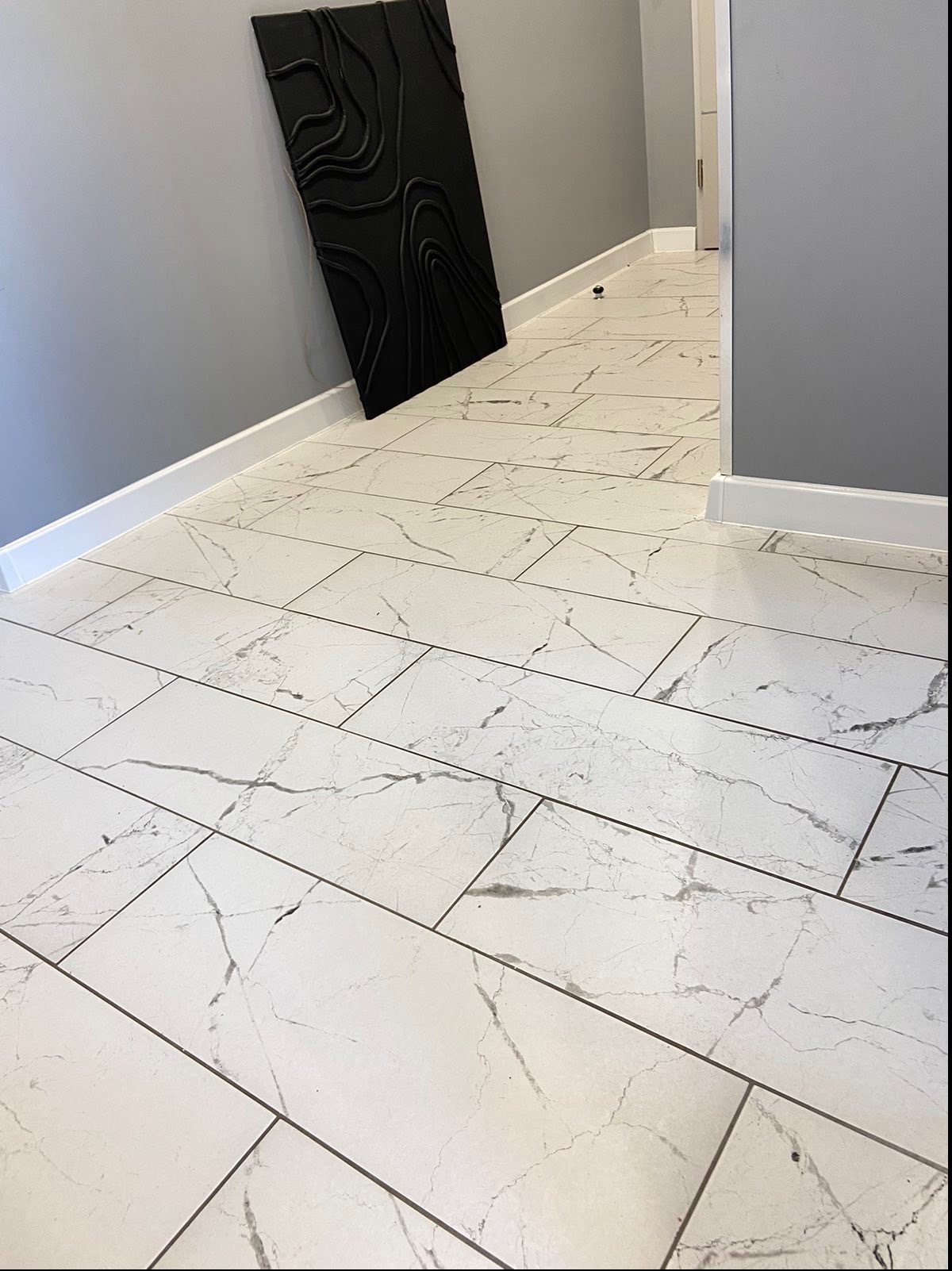 Białe, prostokątne płytki podłogowe z szarym marmurkowym wzorem, ułożone w korytarzu z szarą ścianą i czarną, abstrakcyjną dekoracją ścienną.