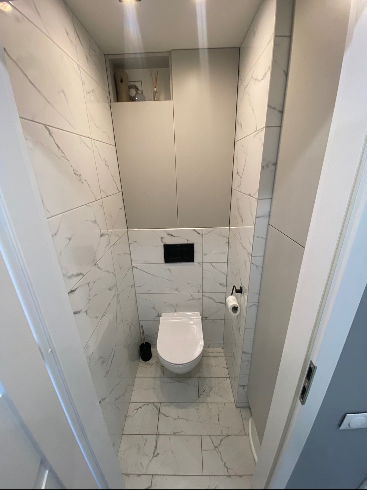 Nowoczesna toaleta w małej łazience, wykończona białymi płytkami imitującymi marmur, z podwieszaną miską WC i zabudową meblową nad nią, oświetlona punktowymi lampami.