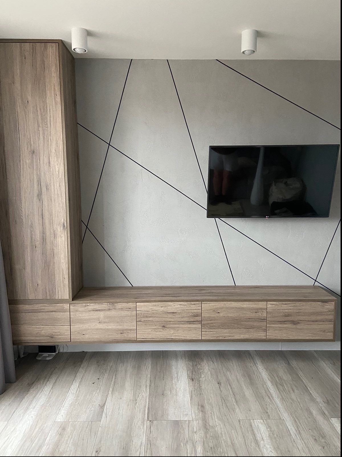 Drewniana zabudowa ściany z szafkami i szafą, telewizor na ścianie z geometrycznym wzorem z czarnych linii, podłoga z paneli w odcieniach szarości i beżu, dwa białe reflektory sufitowe.