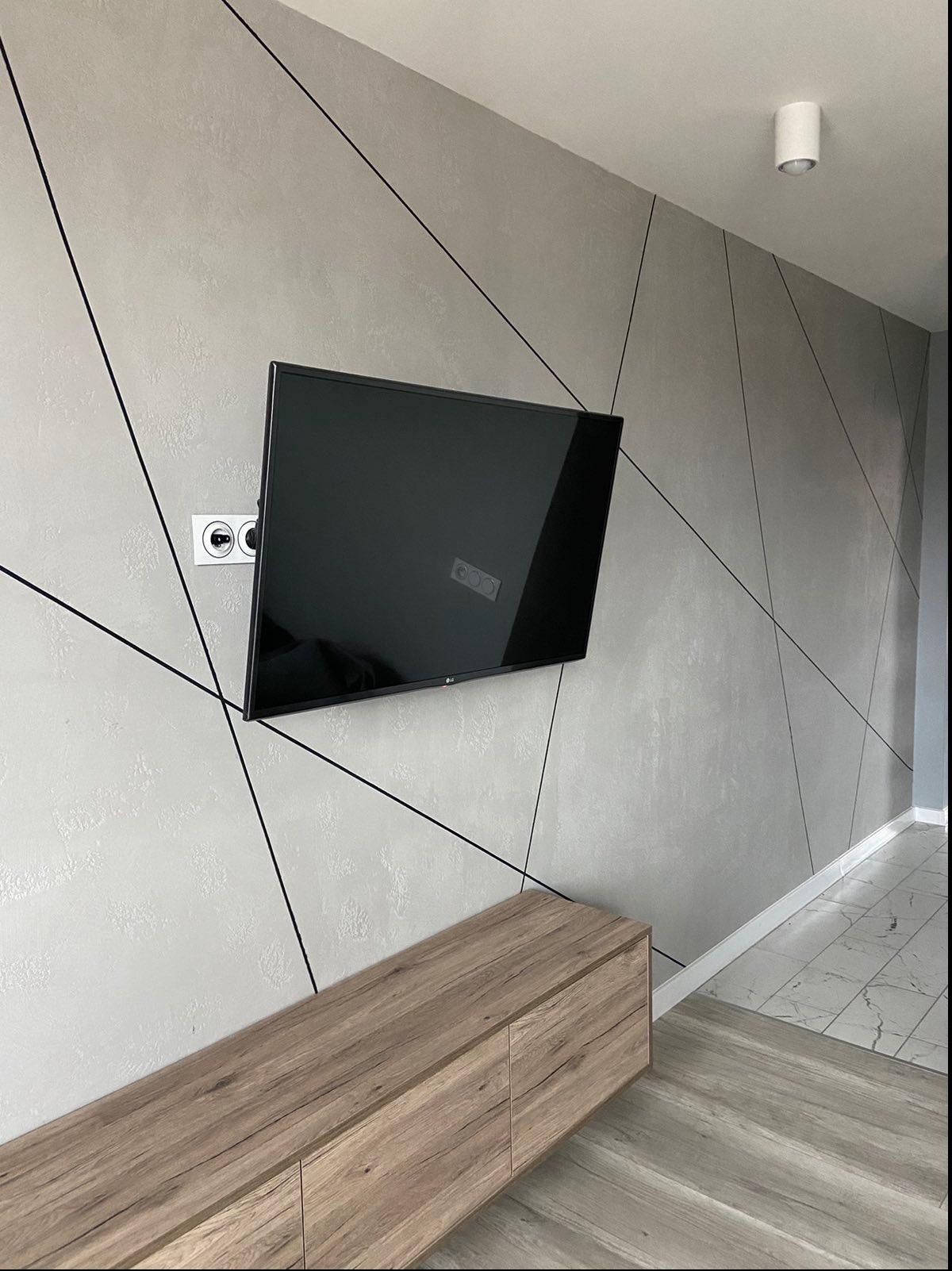 Szara ściana z dekoracyjną strukturą i czarnymi liniami tworzącymi geometryczny wzór, wiszący telewizor, drewniana szafka RTV i białe gniazdko elektryczne.