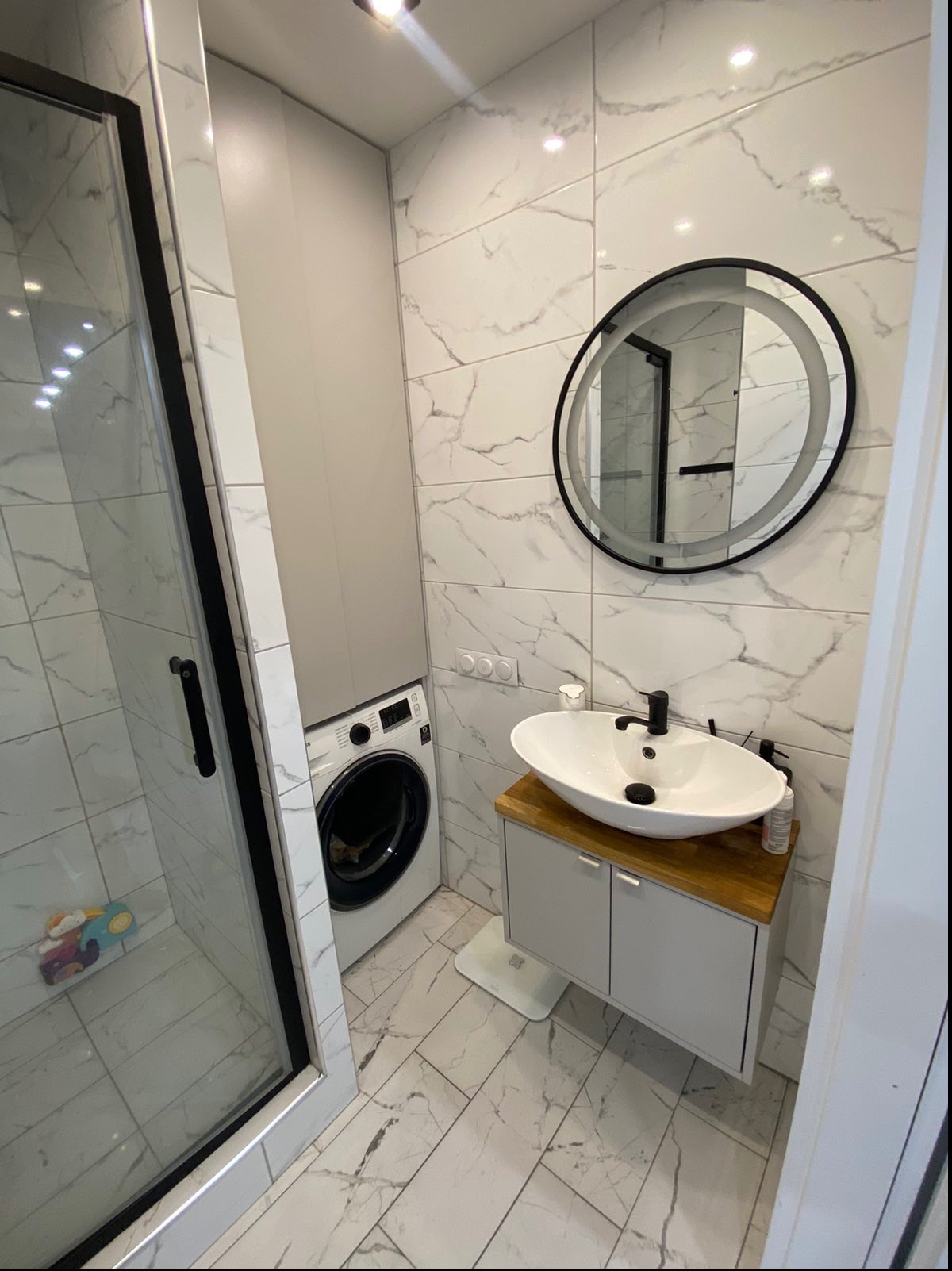 Wykończona łazienka z kabiną prysznicową z czarnymi ramami, pralką zabudowaną w szafce, owalną umywalką na drewnianym blacie i okrągłym lustrem z podświetleniem LED na ścianie w marmurkowe płytki.