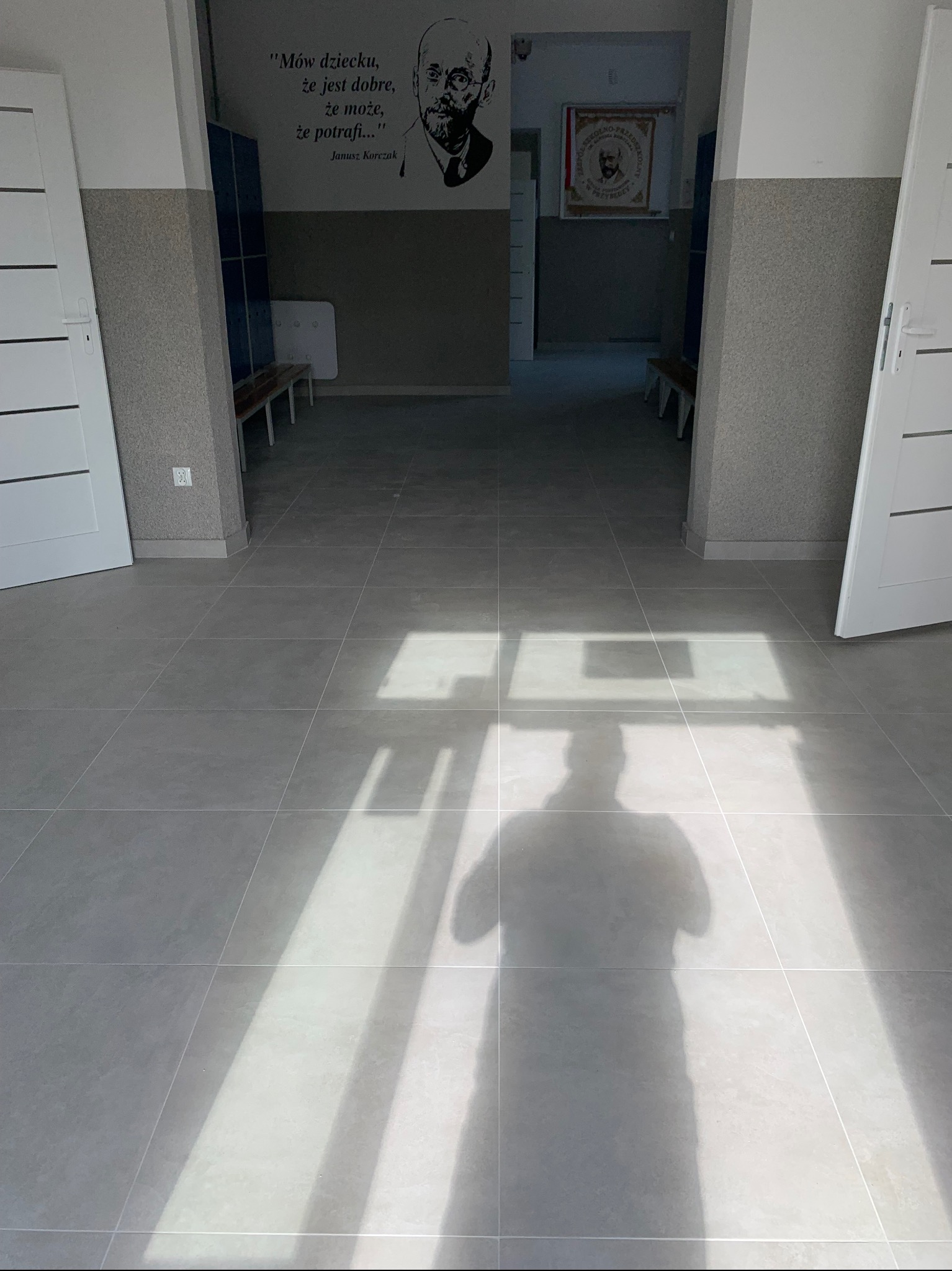 Jasnoszare, kwadratowe płytki podłogowe w korytarzu szkolnym z cieniem fotografa, widoczne drzwi po bokach i portret Janusza Korczaka na ścianie w tle.