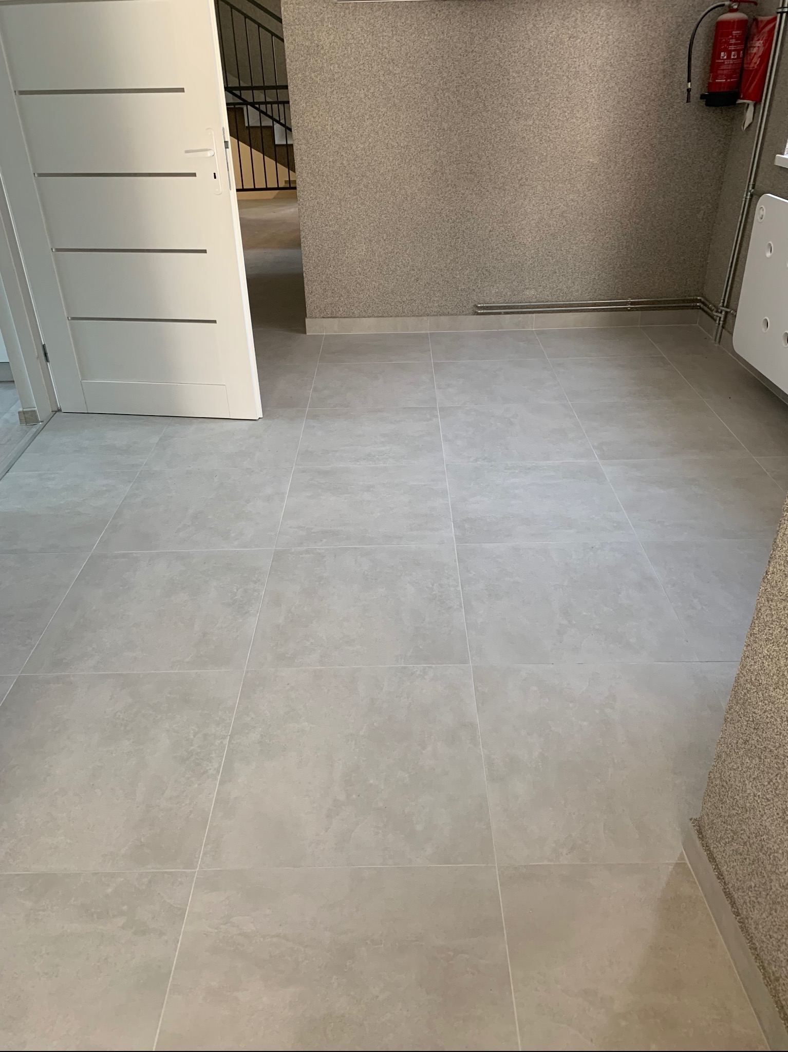 Jasnoszare płytki podłogowe ułożone w kwadraty z wąskimi fugami, widoczna biała ściana z grzejnikiem, czerwona gaśnica, fragment otwartych białych drzwi z poziomymi wgłębieniami i schody w tle.