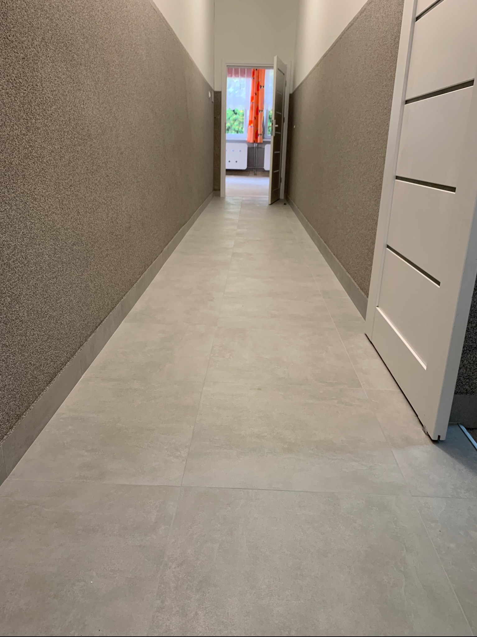 Korytarz po remoncie z jasnymi płytkami podłogowymi, ściany wykończone strukturalnym tynkiem w odcieniach brązu, białe drzwi po prawej i widok na pokój z pomarańczową zasłoną w tle.