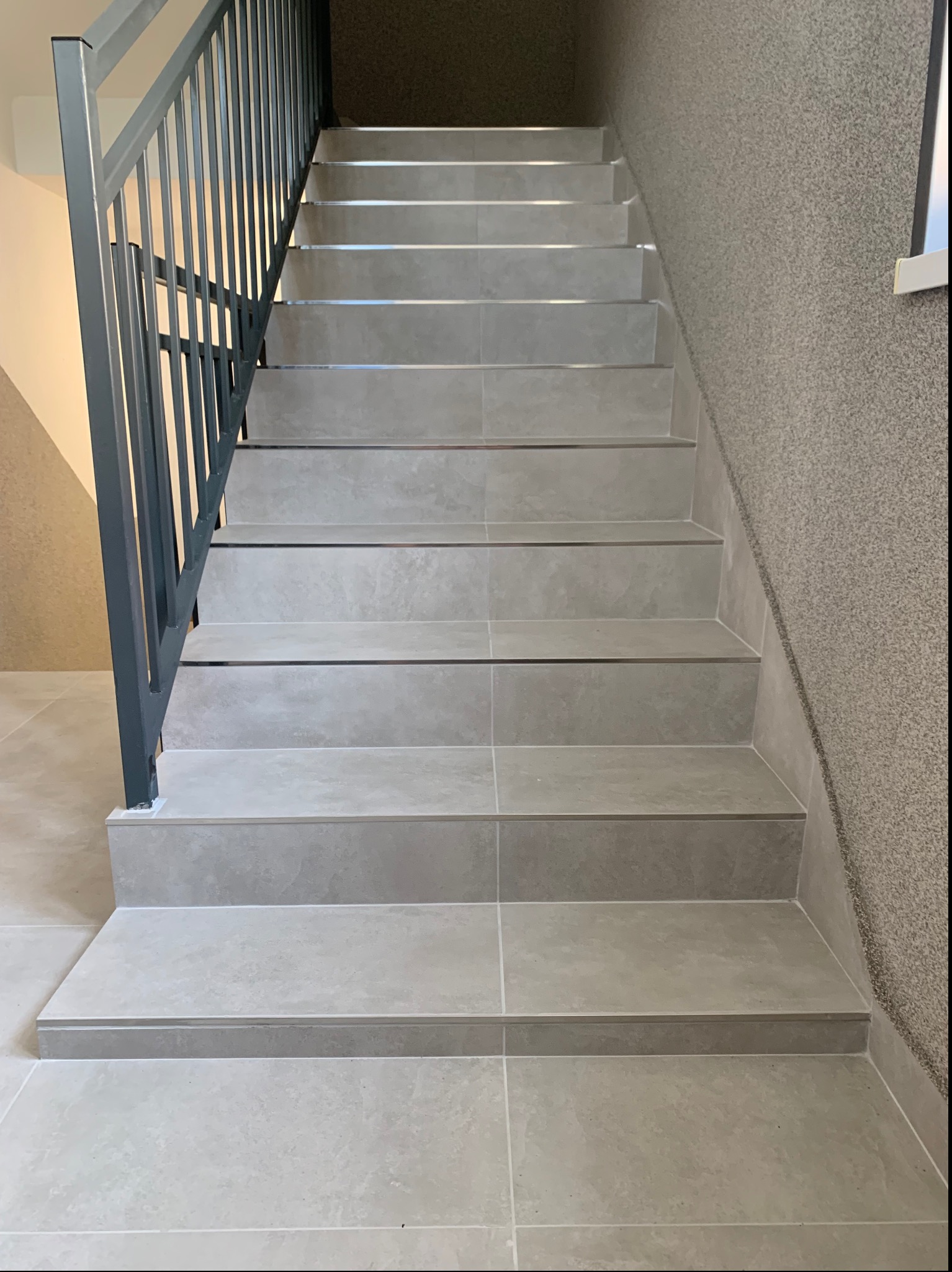 Schody wyłożone szarymi płytkami ceramicznymi z metalowymi listwami zabezpieczającymi krawędzie, widoczne szare ściany o ziarnistej fakturze i metalowa balustrada.