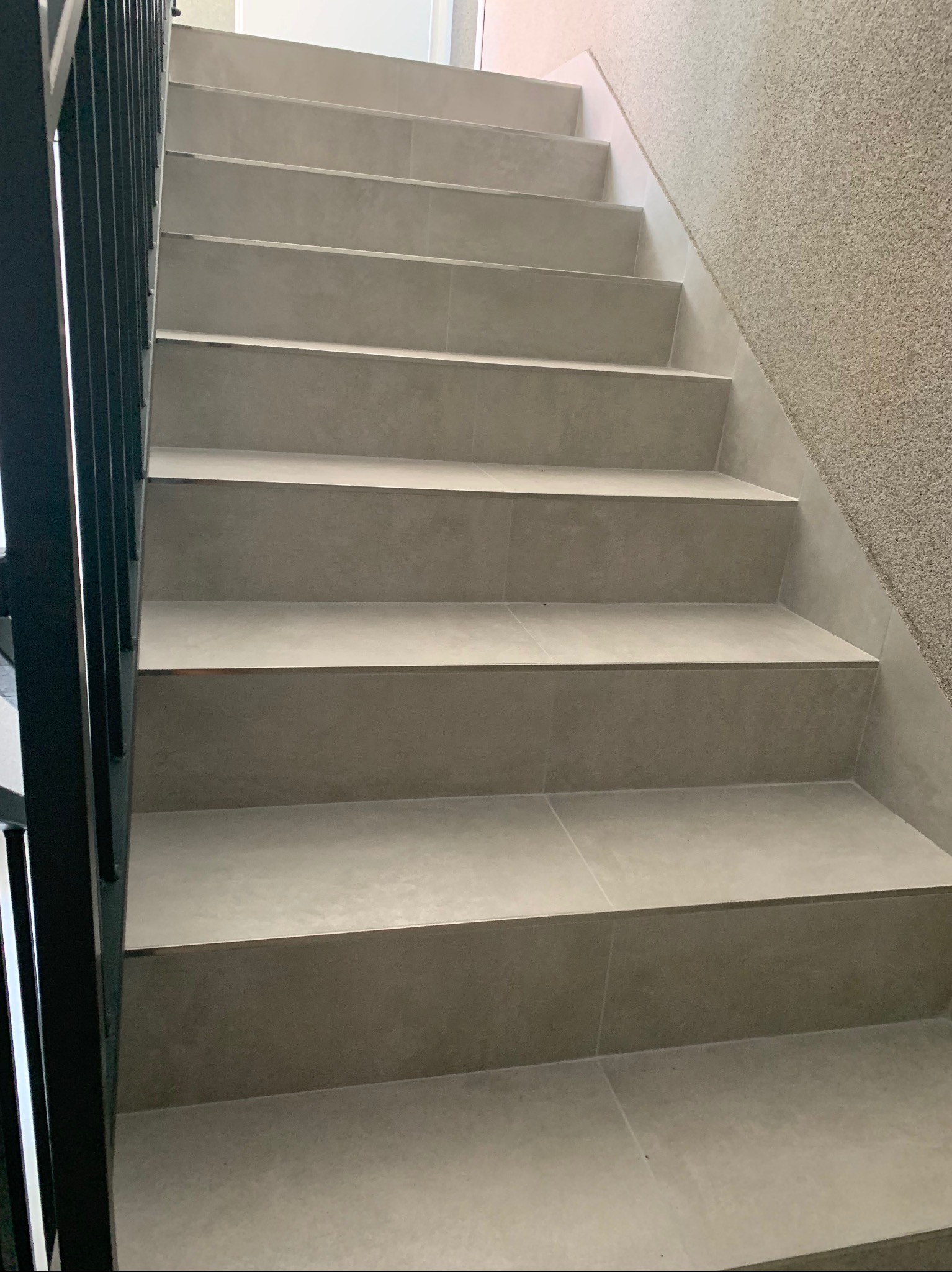 Schody wyłożone szarymi płytkami ceramicznymi, widok z dołu, z czarną metalową barierką po lewej stronie i ścianą o szorstkiej fakturze po prawej.