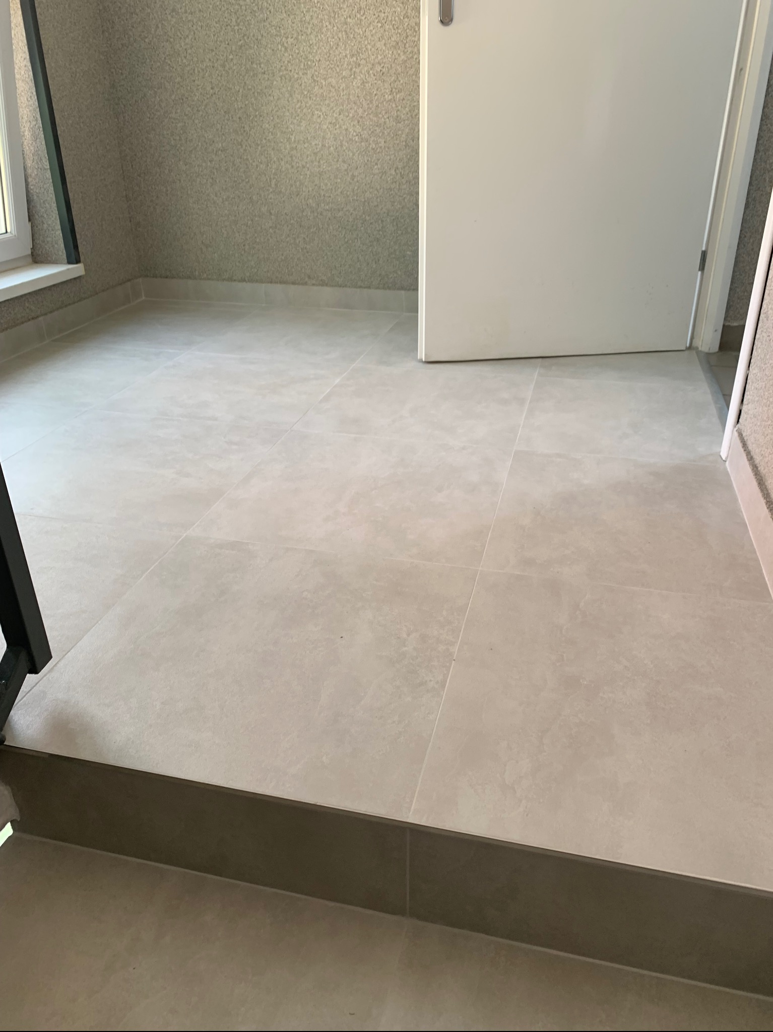 Jasne, kwadratowe płytki podłogowe ułożone w pomieszczeniu z szarą, teksturowaną ścianą i białymi drzwiami.