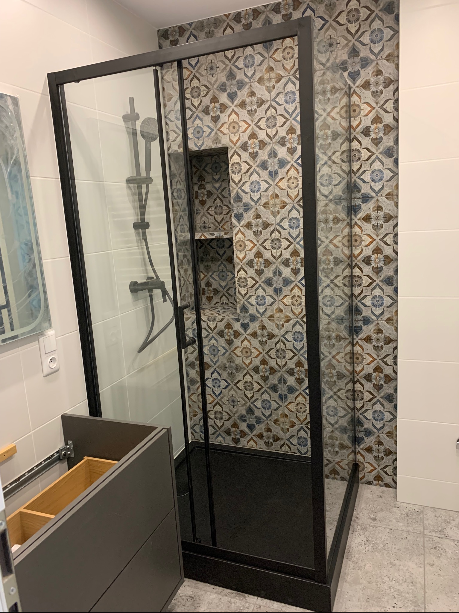 Nowoczesna kabina prysznicowa z czarnymi profilami, wzorzyste płytki na ścianie, niedokończona szafka pod umywalkę z wysuwanymi szufladami.