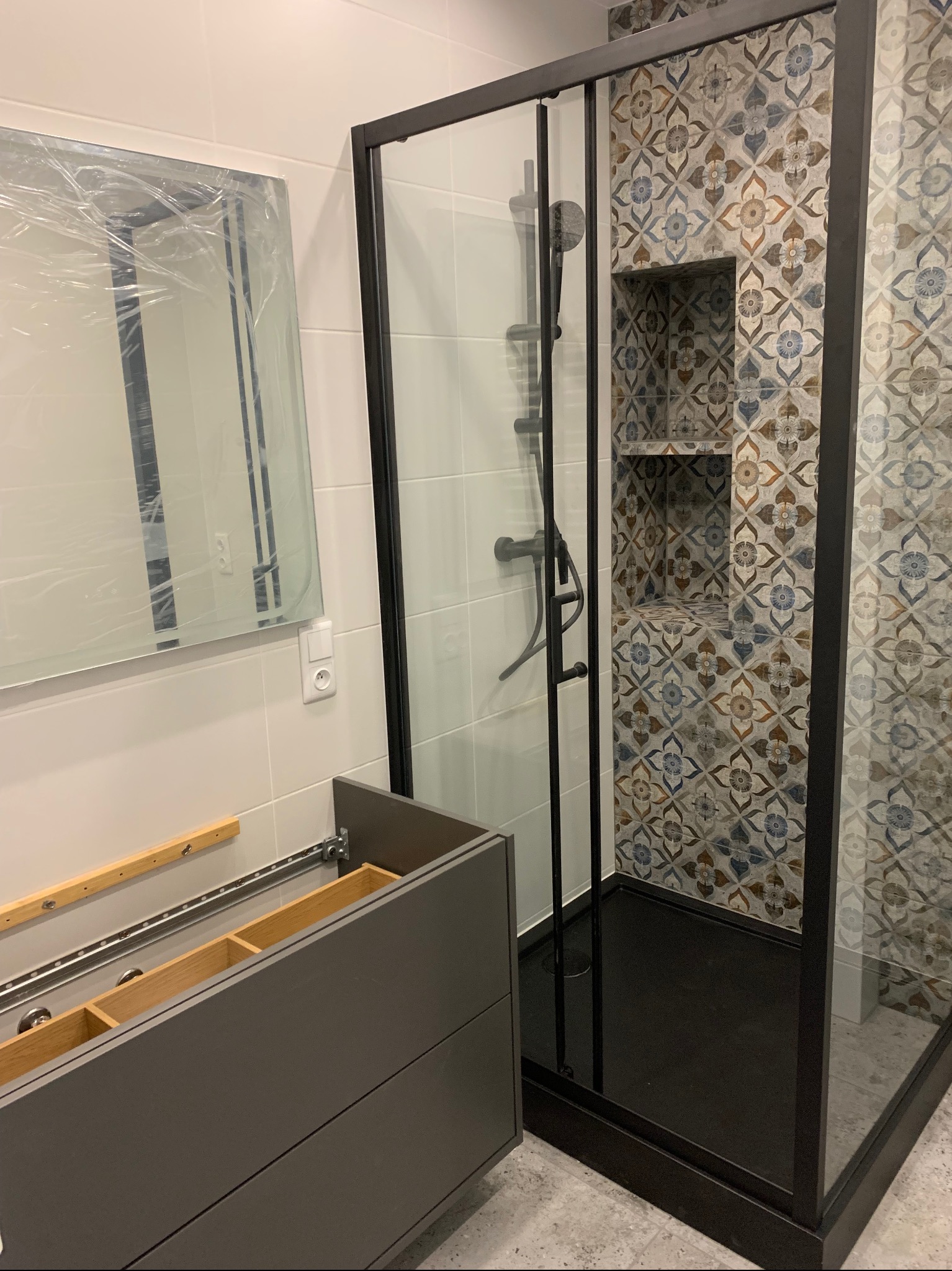 Montaż kabiny prysznicowej z czarną ramą i wzorzystymi płytkami wnękowymi, lustro w folii ochronnej i niedokończona szafka pod umywalkę.