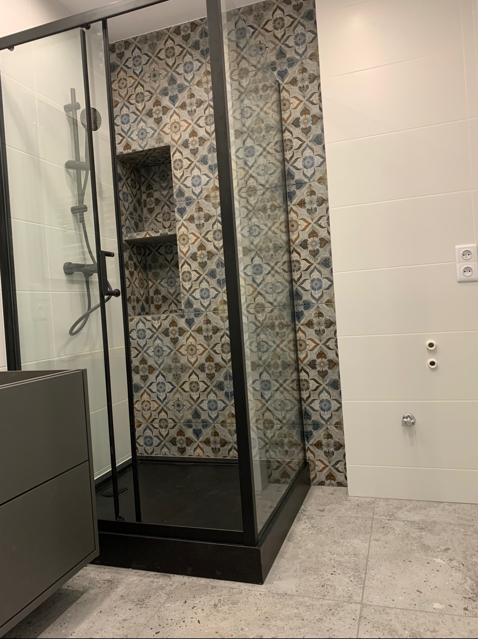 Nowoczesna kabina prysznicowa z czarną ramą, wyłożona płytkami w geometryczne wzory, z wnęką na kosmetyki, sąsiadująca z białą ścianą z poziomymi panelami i gniazdkiem elektrycznym.