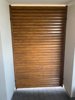 Drewnopodobna, pozioma przesłona z listew zamontowana pomiędzy dwoma białymi ścianami na zewnątrz budynku.