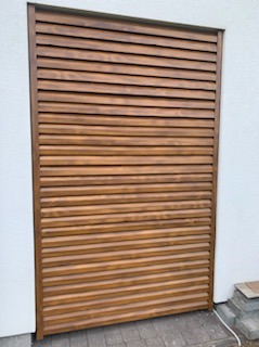 Drewniana przesłona lamelowa w kolorze brązowym, zamontowana na białej ścianie, widoczna kostka brukowa u podstawy.