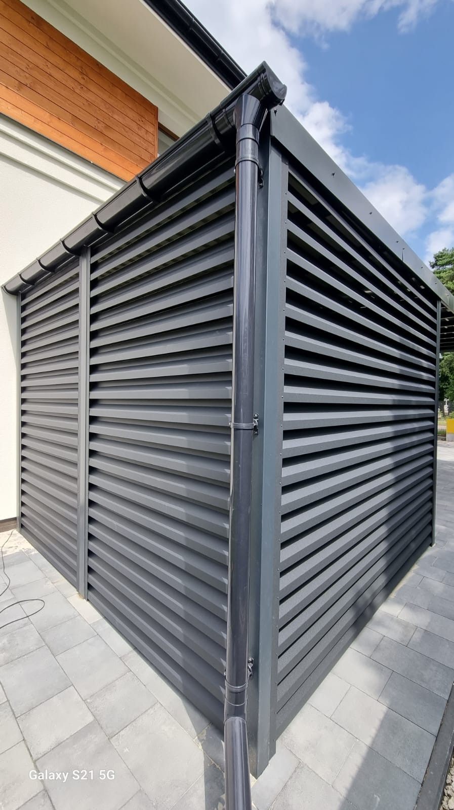 Szara pergola z metalowymi lamelami, zintegrowana z budynkiem, widoczny system rynnowy i kostka brukowa.