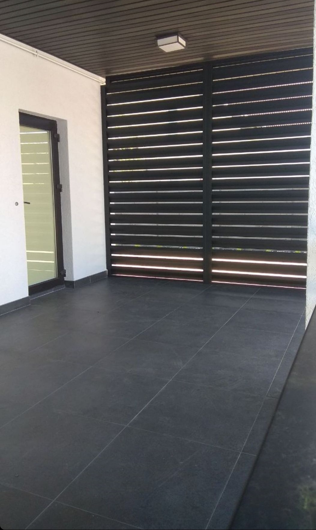 Nowoczesna, pozioma, czarna przesłona z metalowych listew, zamontowana na tarasie z ciemnej terakoty, przylegająca do białej ściany budynku z przeszklonymi drzwiami.