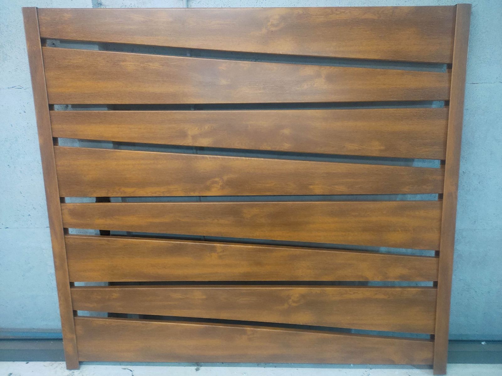 Panel ogrodzeniowy z poziomych, ukośnie ułożonych desek w odcieniach brązu, widoczny na tle szarej ściany.