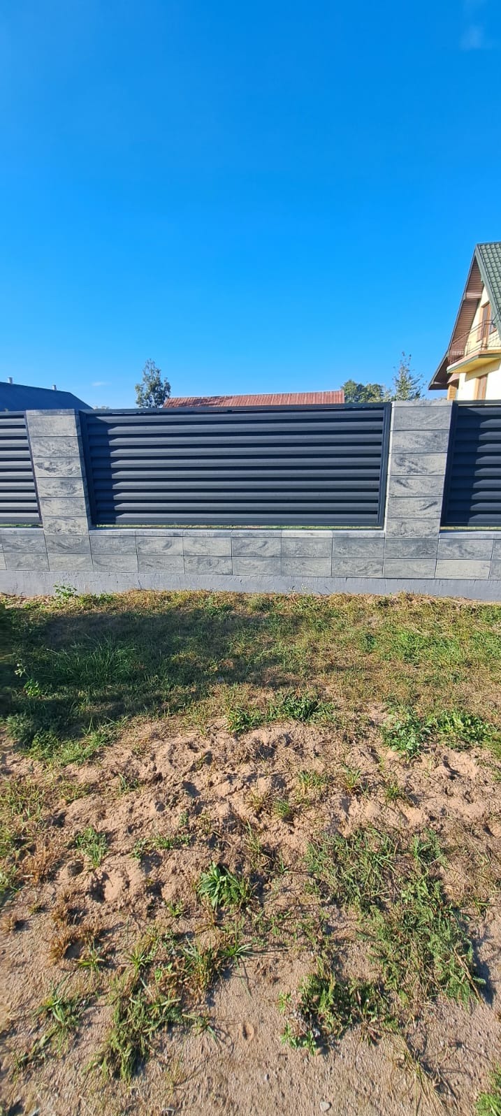 Nowoczesne ogrodzenie z poziomych, grafitowych paneli, osadzone na betonowej podmurówce i słupkach z szarego betonu architektonicznego, na tle błękitnego nieba i fragmentu domu.