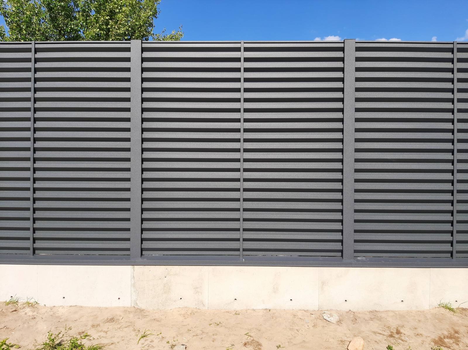 Nowoczesne, poziome panele w kolorze grafitowym tworzące ogrodzenie na betonowej podmurówce, widoczne drzewa i błękitne niebo w tle.