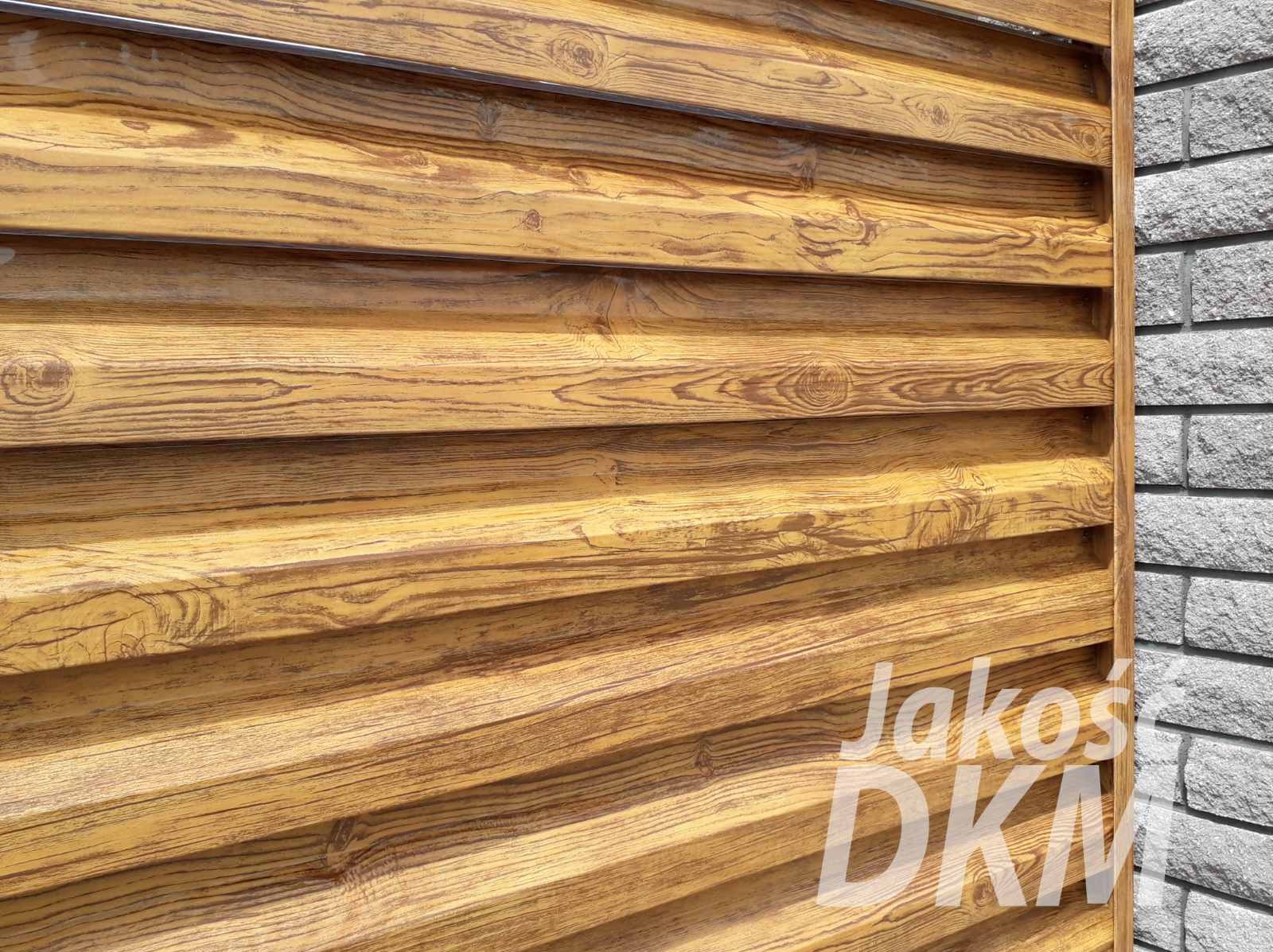 Bliski kadr fragmentu brązowego ogrodzenia panelowego z imitacją drewna, z widocznym logo firmy Jakos DKM w prawym dolnym rogu i krawędzią muru z szarej cegły.