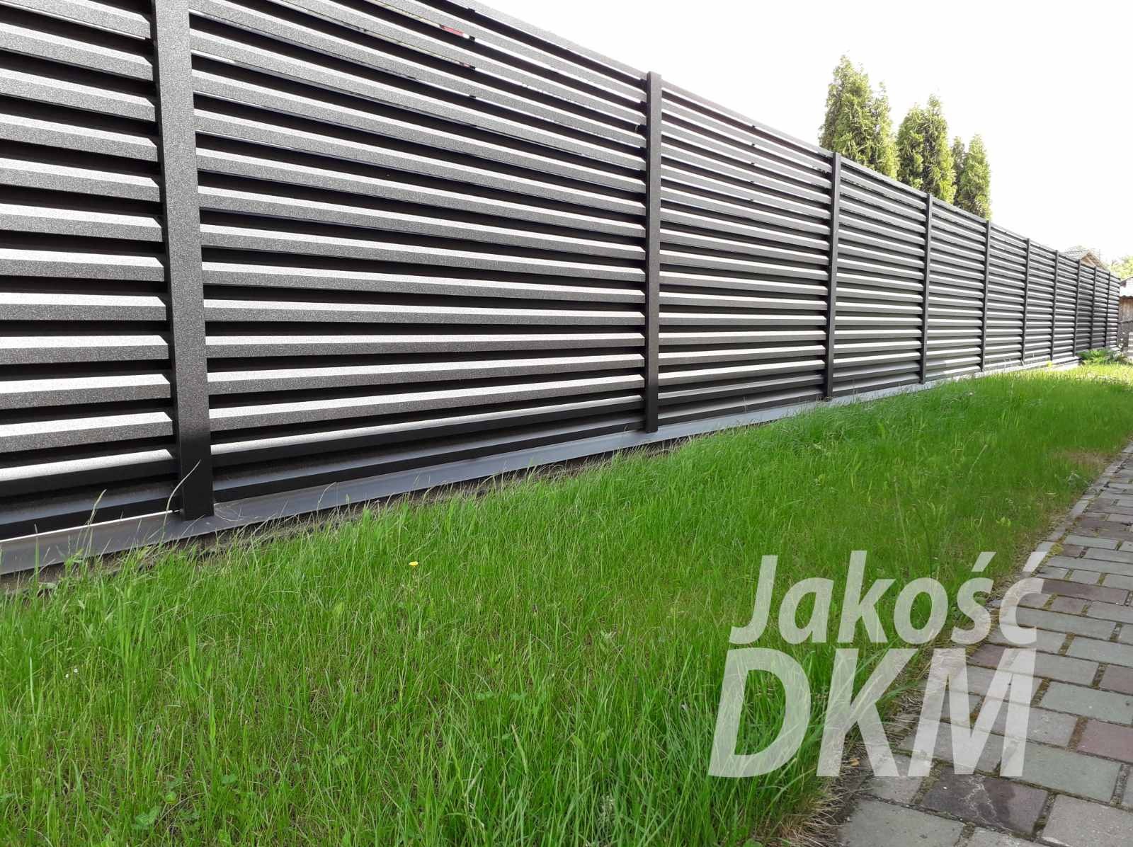 Długie, poziome przęsła w kolorze grafitowym tworzą nowoczesne ogrodzenie posesji, widoczne z poziomu trawnika i chodnika z kostki.